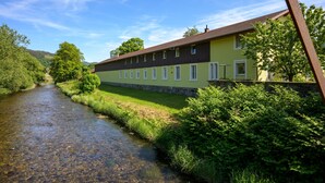 A building with a long side, a river in front, and green trees around. - La Maison du Portier: Mountain View, Private Terrace with Fire Pit and Wi-Fi (Malmerspach)
