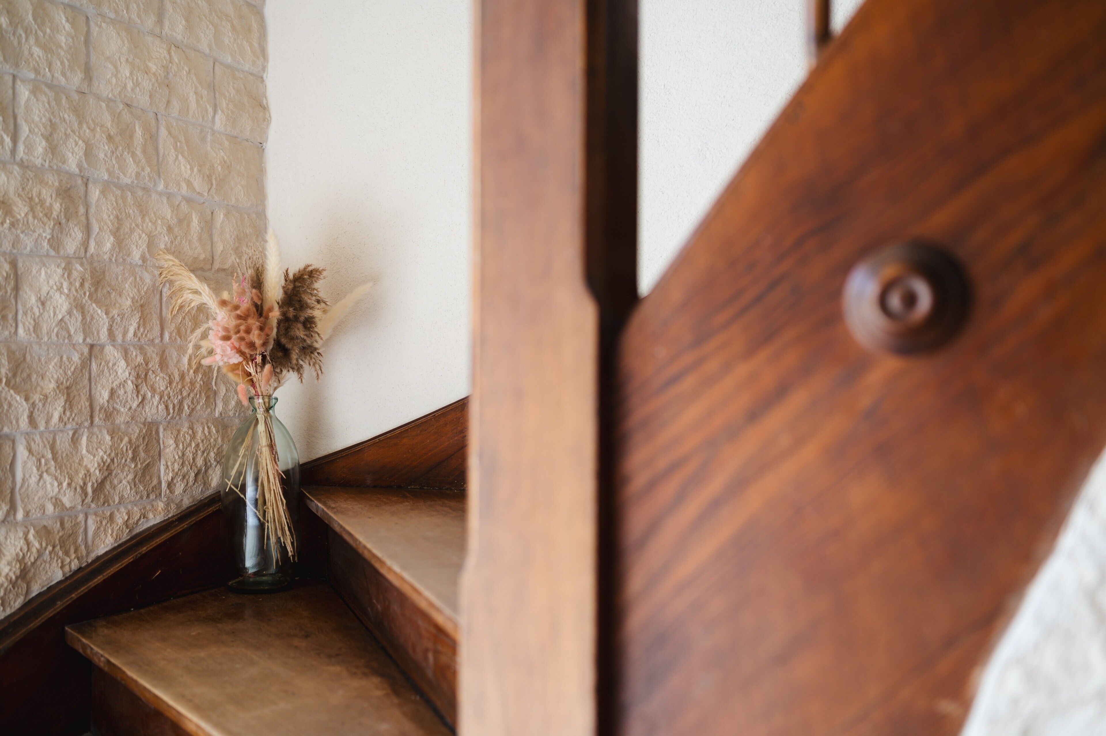 Un escalier en bois, et sur le palier, un vase en verre contenant des fleurs séchées.