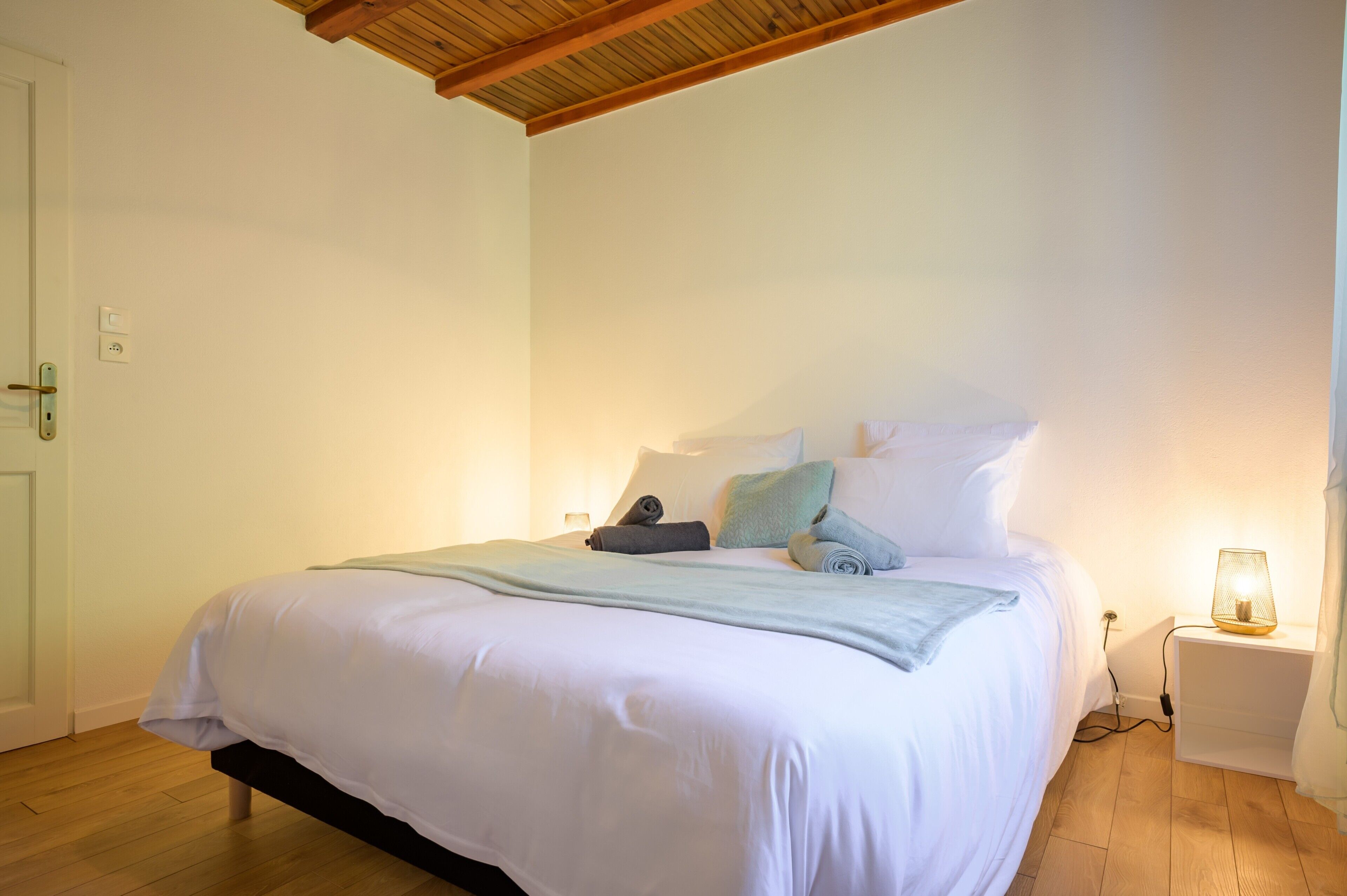 A neatly made bed with a white comforter and pillows, a bedside lamp, and a wooden floor.