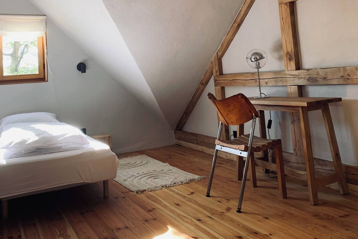 Un dormitorio con techo inclinado, vigas de madera, una cama, un escritorio y una silla.