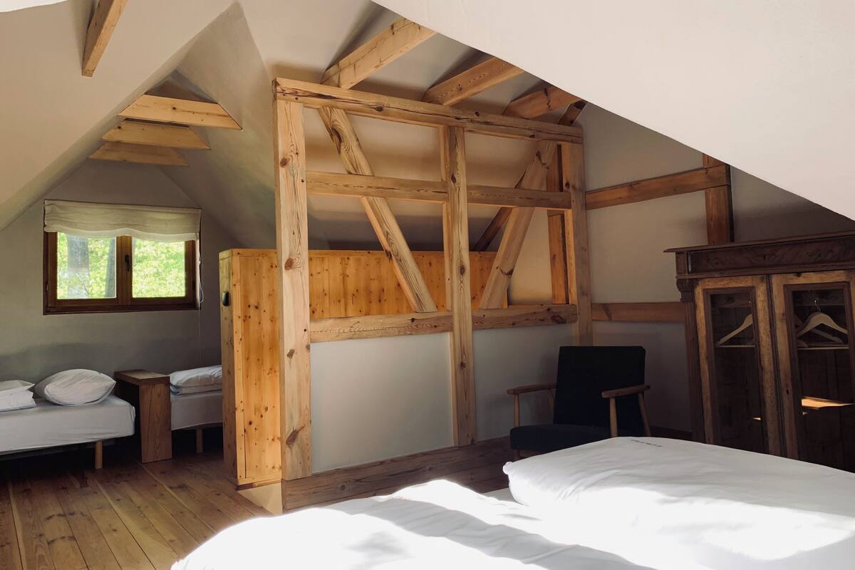 Una habitación con techo inclinado, vigas de madera, una cama, una silla, una mesita y un armario con puertas de cristal.