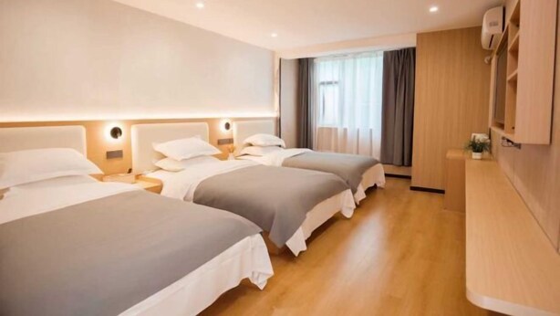 Habitación de hotel con tres camas, piso de madera, mesita de noche con lámpara, ventana con cortinas y unidad de aire acondicionado de pared.