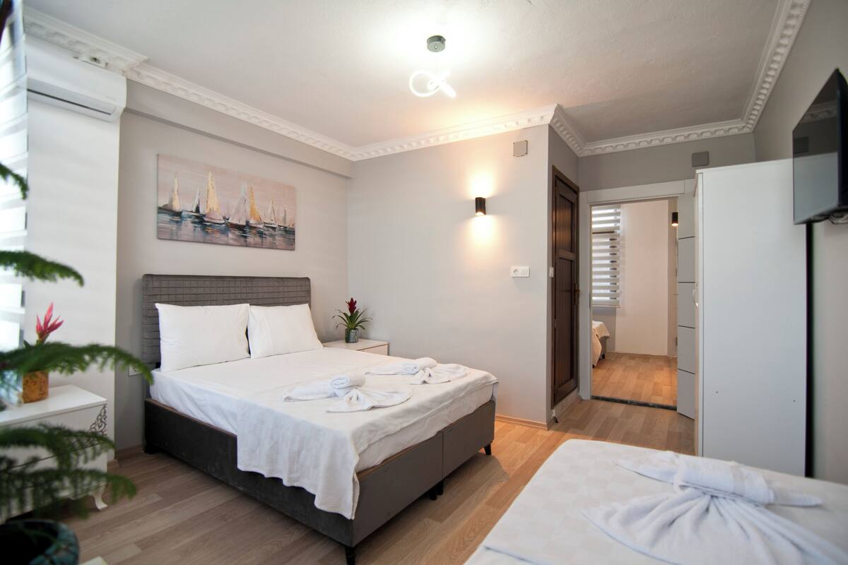 A hotel room with a bed, a bedside table, a wall-mounted light, a painting, and a door leading to another room.
