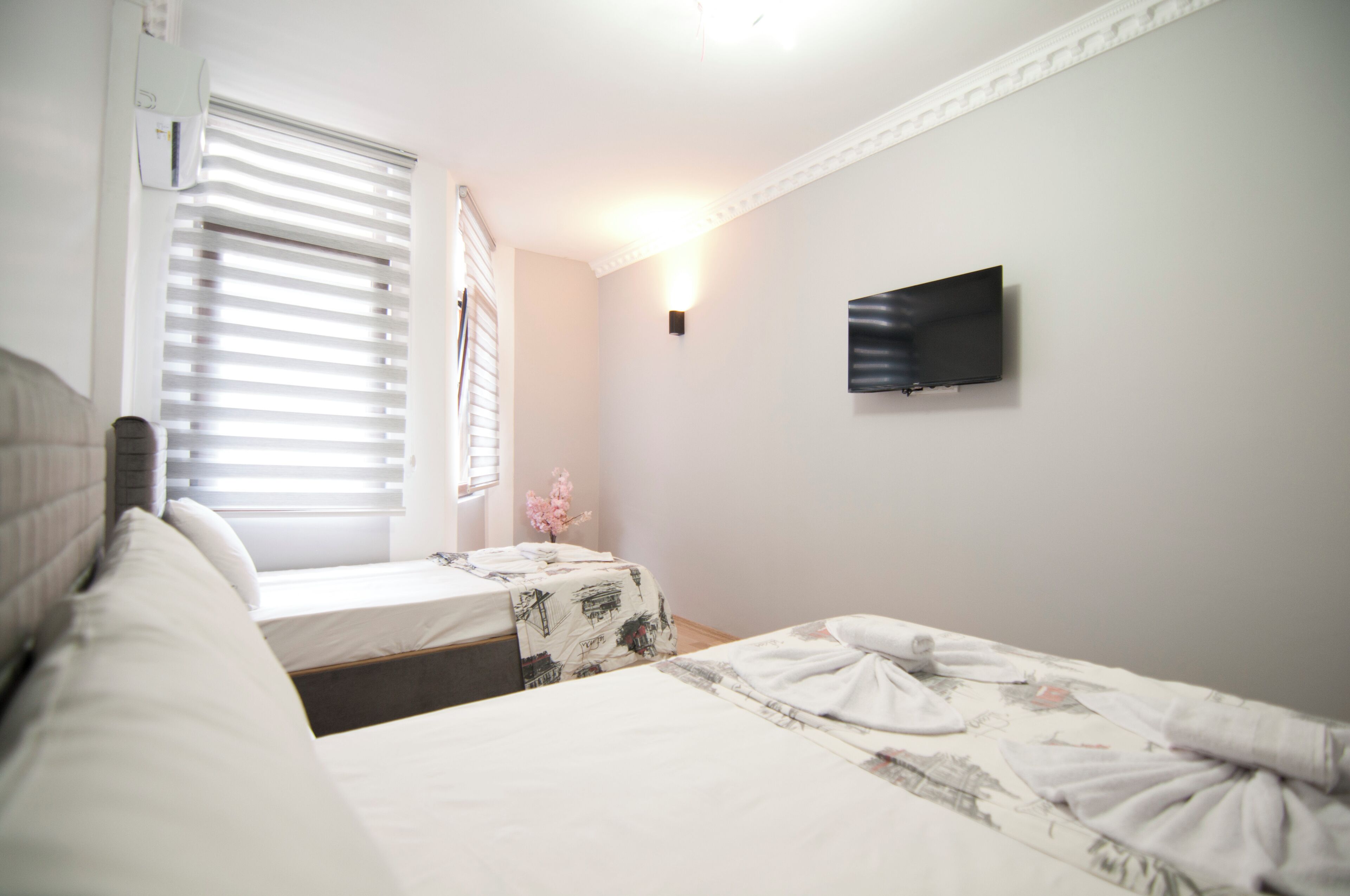 A bedroom with two beds, a TV mounted on the wall, and a window with blinds.