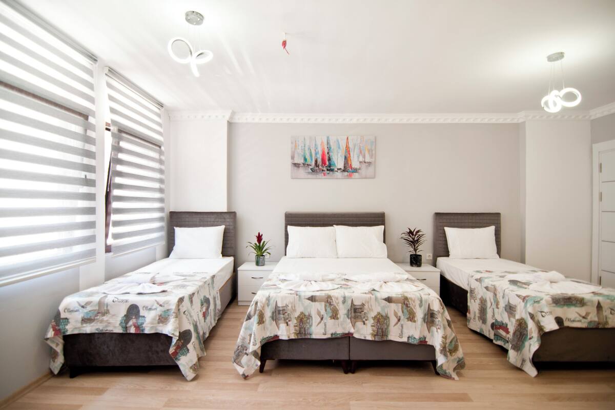 A hotel room with three single beds, a painting on the wall, and a window with blinds.