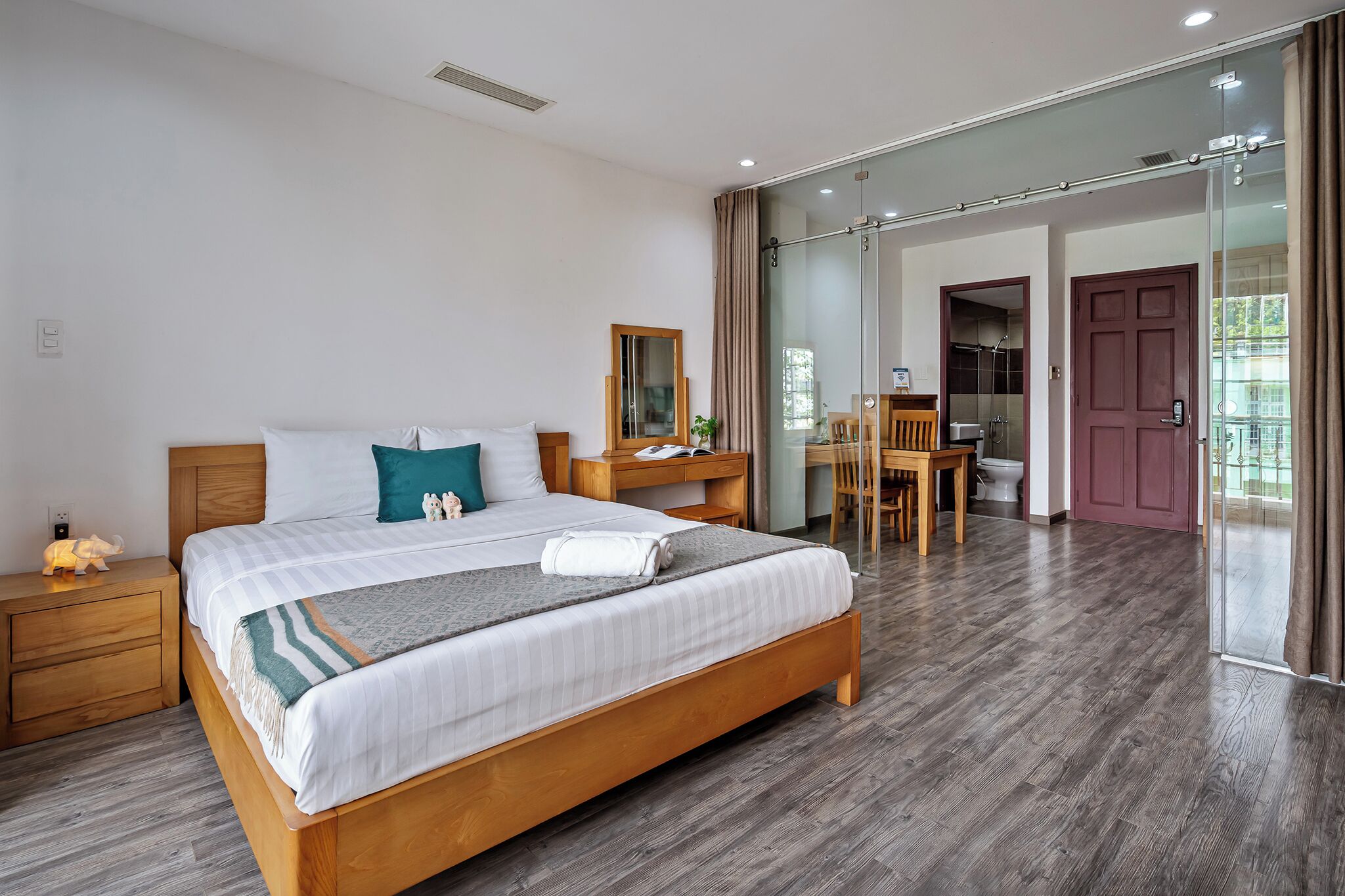 A modern hotel room with a wooden bed, a desk, a chair, and a glass door leading to a balcony.