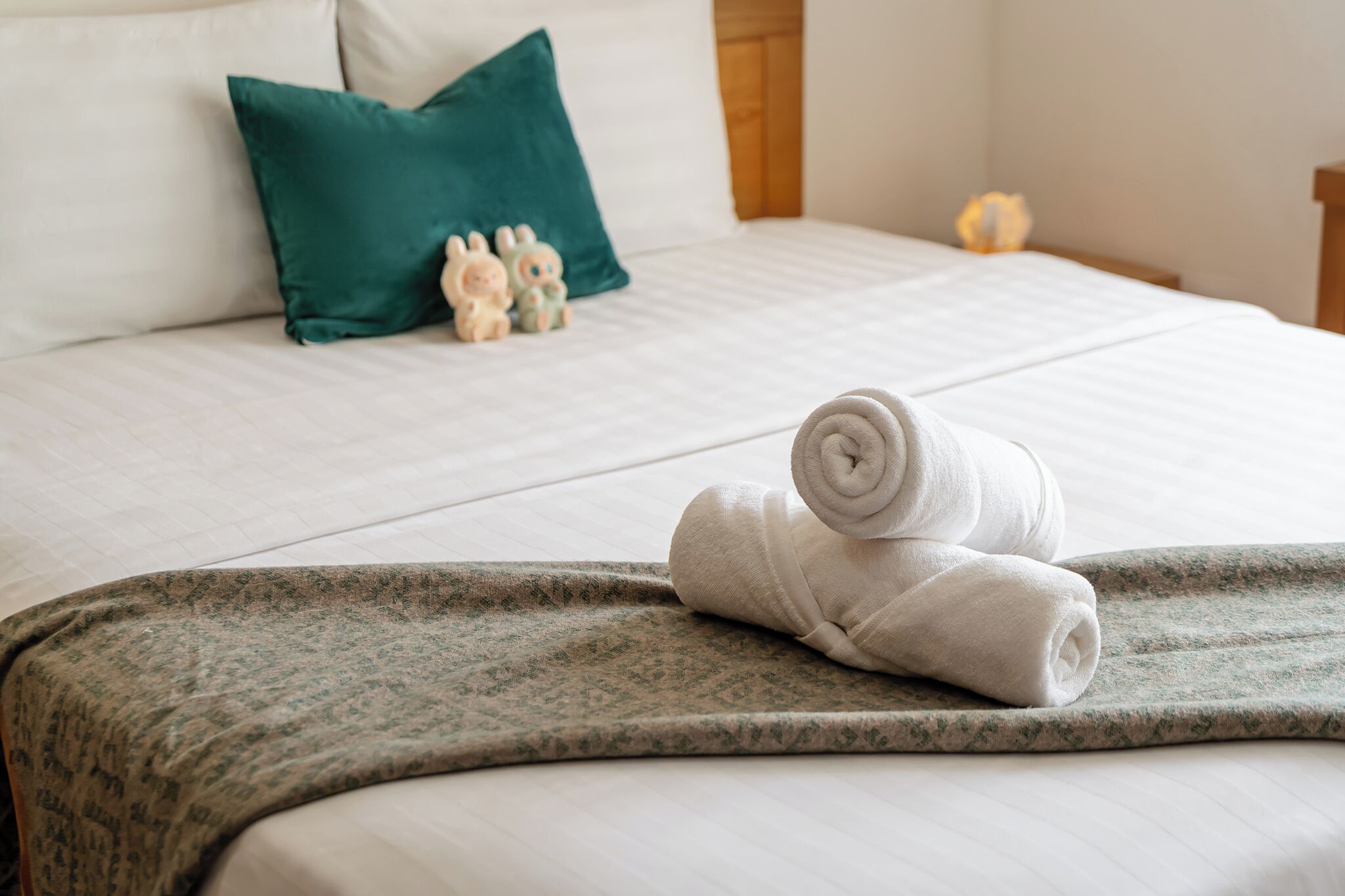 Una cama bien hecha con una toalla blanca enrollada, un cojín verde y una pequeña figurita.