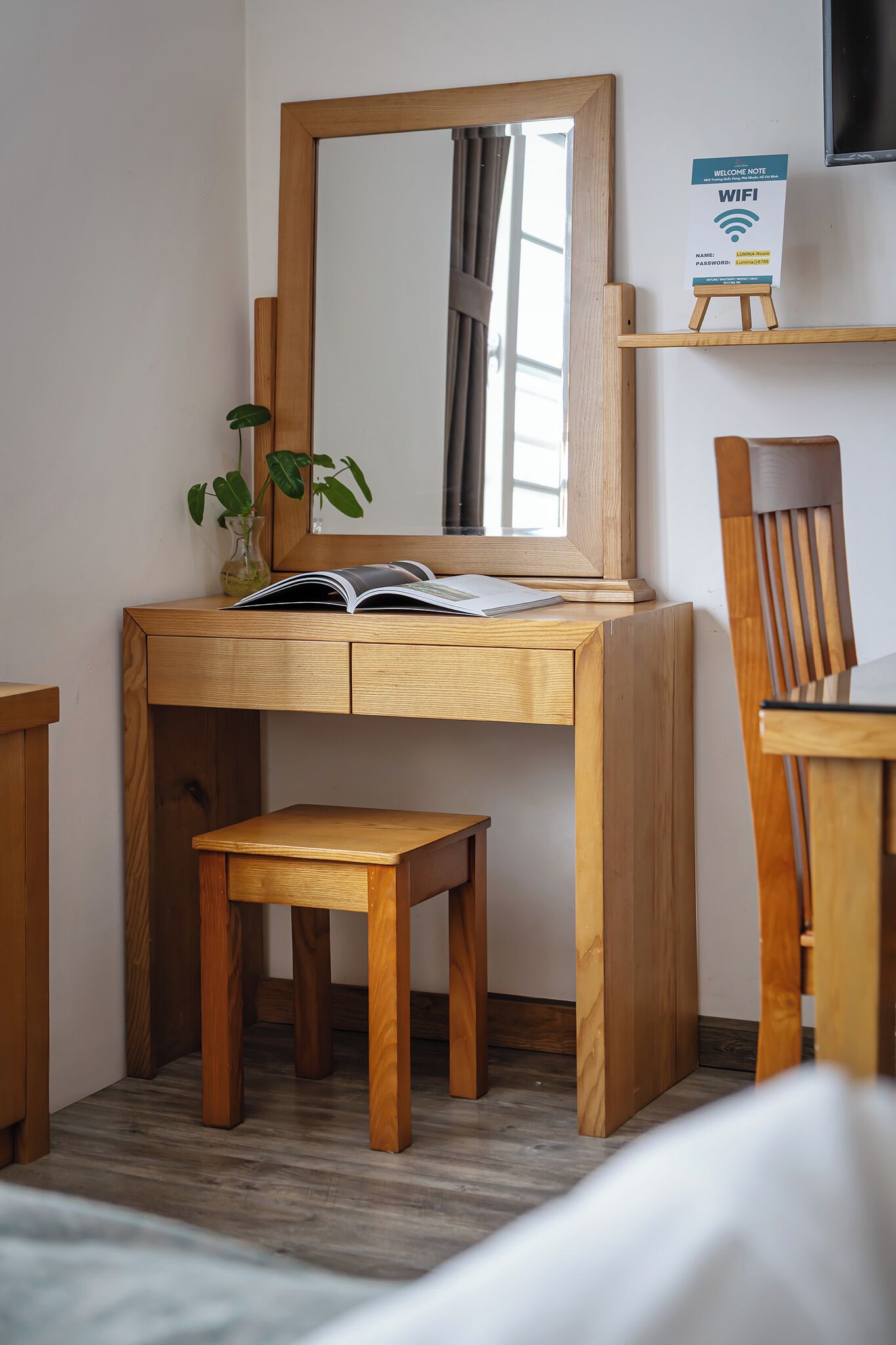 A wooden desk with a mirror, a small stool, a book, a vase with a plant, and a sign indicating WiFi availability.