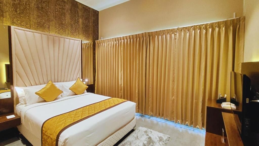A hotel room with a large bed, a headboard, a curtain, a nightstand, and a television.