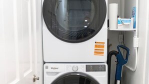 Een kleine wasruimte met twee Whirlpool wasmachines en drogers boven elkaar, een blauwe stofzuiger en een klein plankje met een zak zakdoekjes.