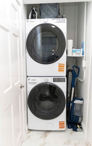 A compact laundry room with two stacked Whirlpool washers and dryers, a blue vacuum cleaner, and a small shelf with a box of tissues. - Chic 3BR in Flatiron/Chelsea | W/D + Smart TV’s (New York)