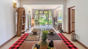 A hallway with a checkered floor leading to a dining area with a table set for two, a large wooden sculpture, and a view of a garden through glass doors.
