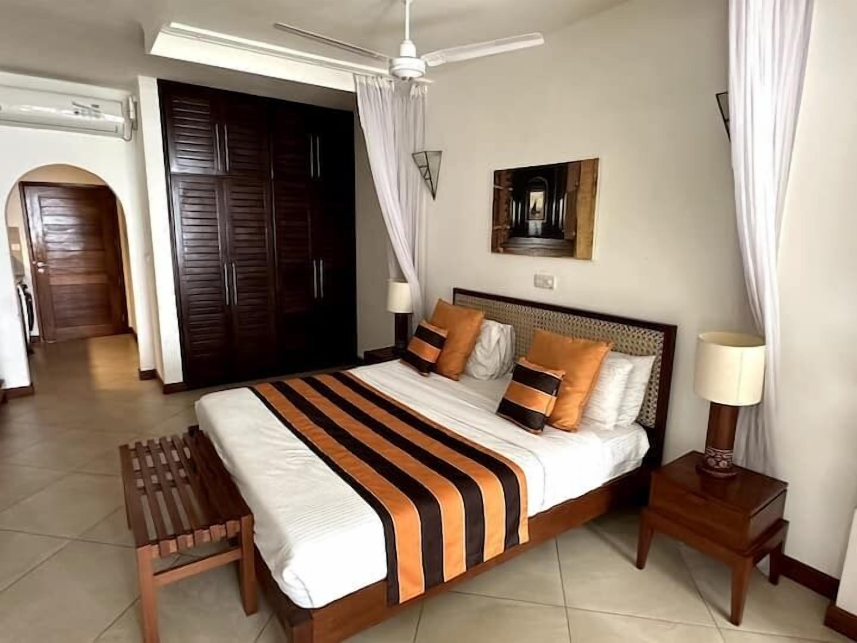 Ein Schlafzimmer mit Bett, Nachttischen, Deckenventilator und einer Tür zu einem weiteren Raum.