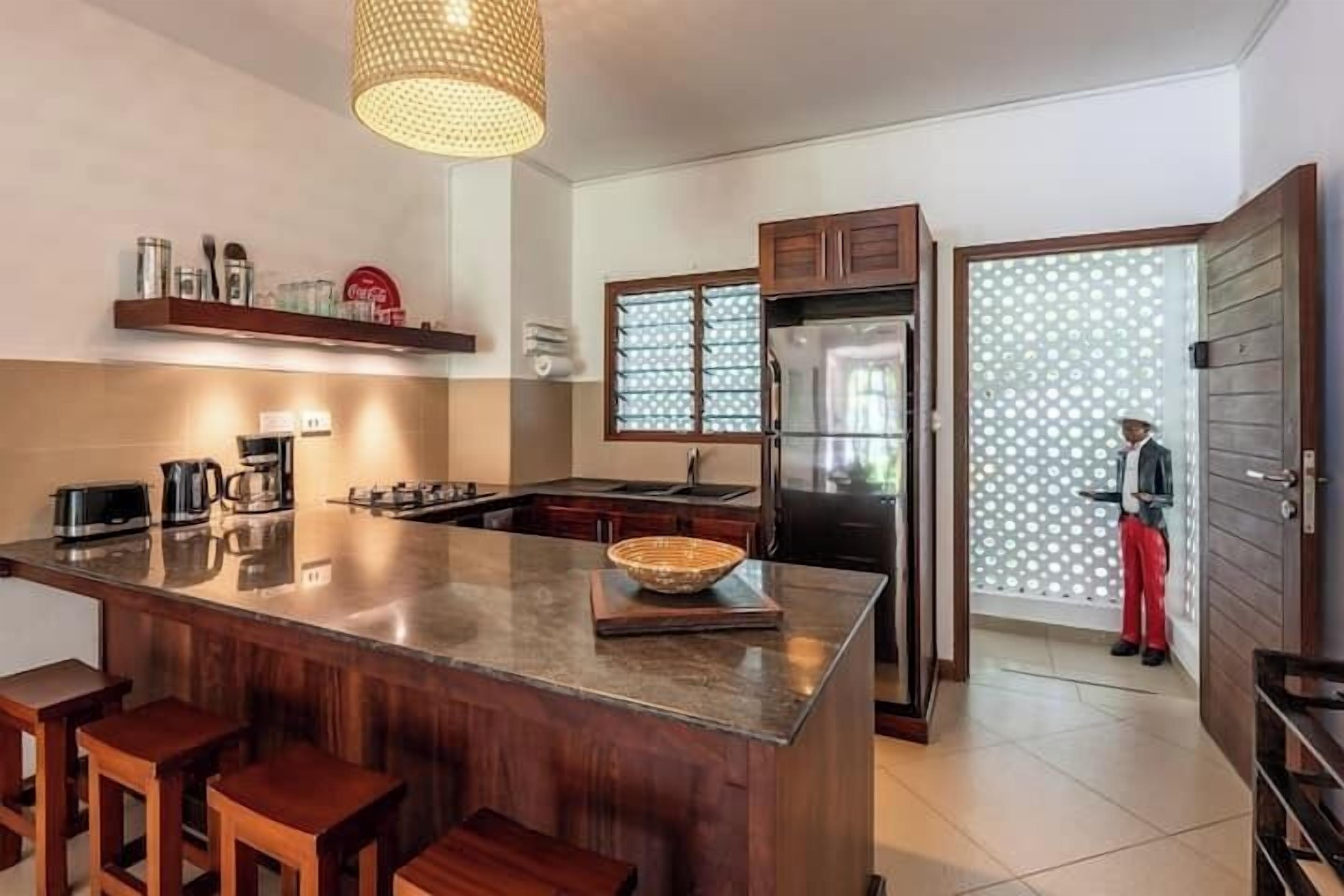 A kitchen with a central island, bar stools, a refrigerator, and a person in red pants standing by the door.
