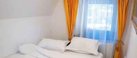 Una cama individual con sábanas blancas, una toalla enrollada y una ventana con cortinas amarillas.