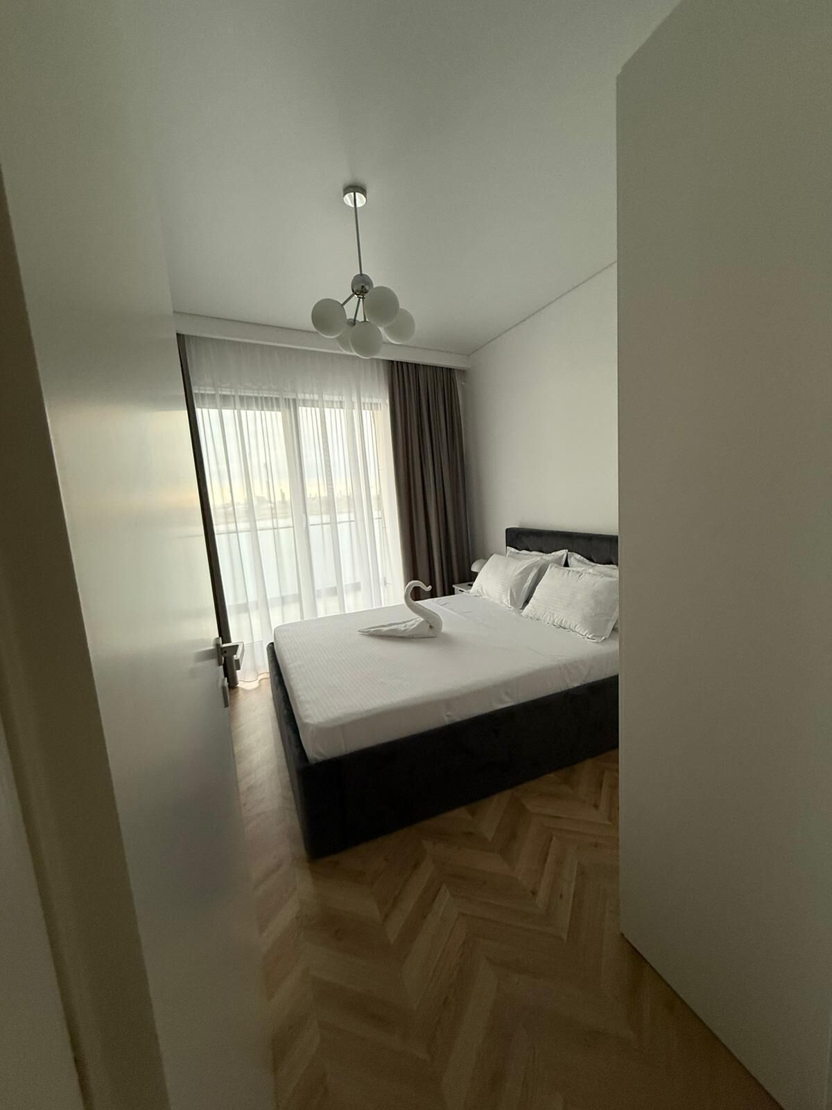 Une chambre à coucher avec un lit, un luminaire au plafond, une fenêtre avec des rideaux et un plancher en bois.