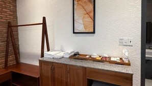 Habitación de hotel moderna con tocador de madera, un refrigerador pequeño, aire acondicionado de pared y una obra de arte con hojas enmarcada.