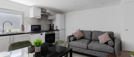 A modern kitchen with white cabinets, a dining area with a black table, and a grey sofa with cushions.
