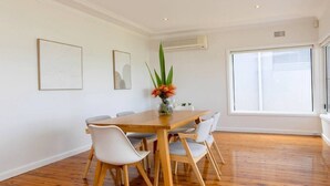 Un coin repas avec une table en bois et des chaises blanches, un vase rempli de fleurs et une grande fenêtre avec des stores.