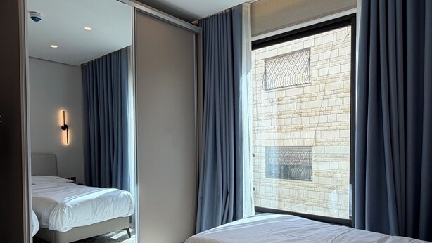 Habitación de hotel con dos camas, un espejo grande, una ventana con cortinas y una pared de piedra visible a través de la ventana.