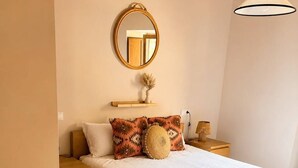 A bedroom with a bed, bedside tables, a mirror, a hanging light fixture, and a wooden floor.