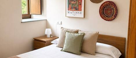 Un dormitorio con cama, mesita de noche, lámpara, ventana y adornos en la pared, incluyendo un cuadro enmarco y dos posavasos.