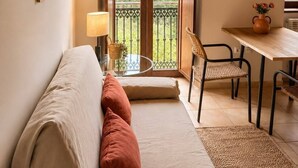 A cozy room with a wooden ceiling, a sofa with cushions, a table with chairs, and a balcony with a view of the forest.