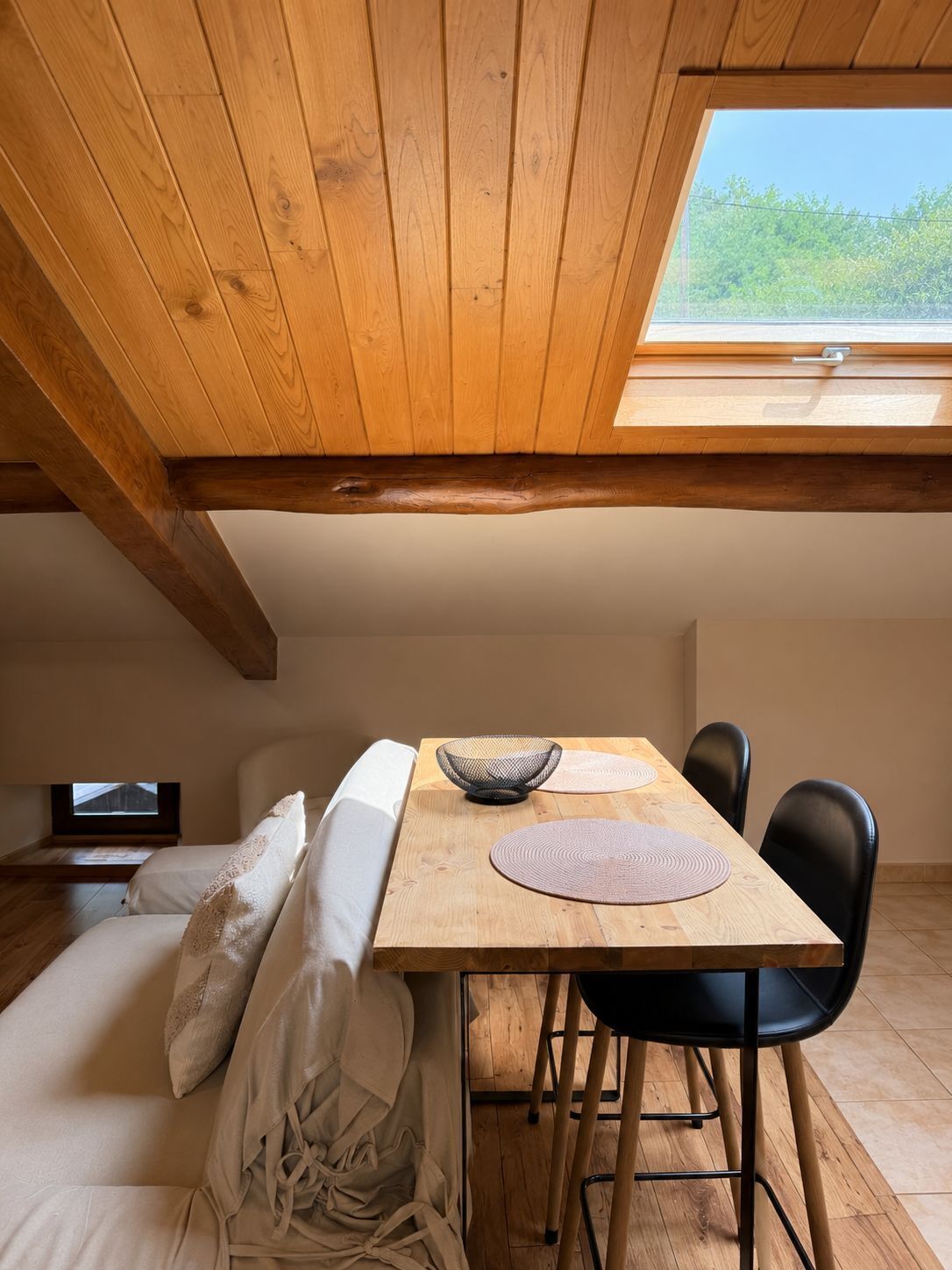 A cozy room with a wooden ceiling, a skylight, a wooden dining table with chairs, and a bed with pillows.