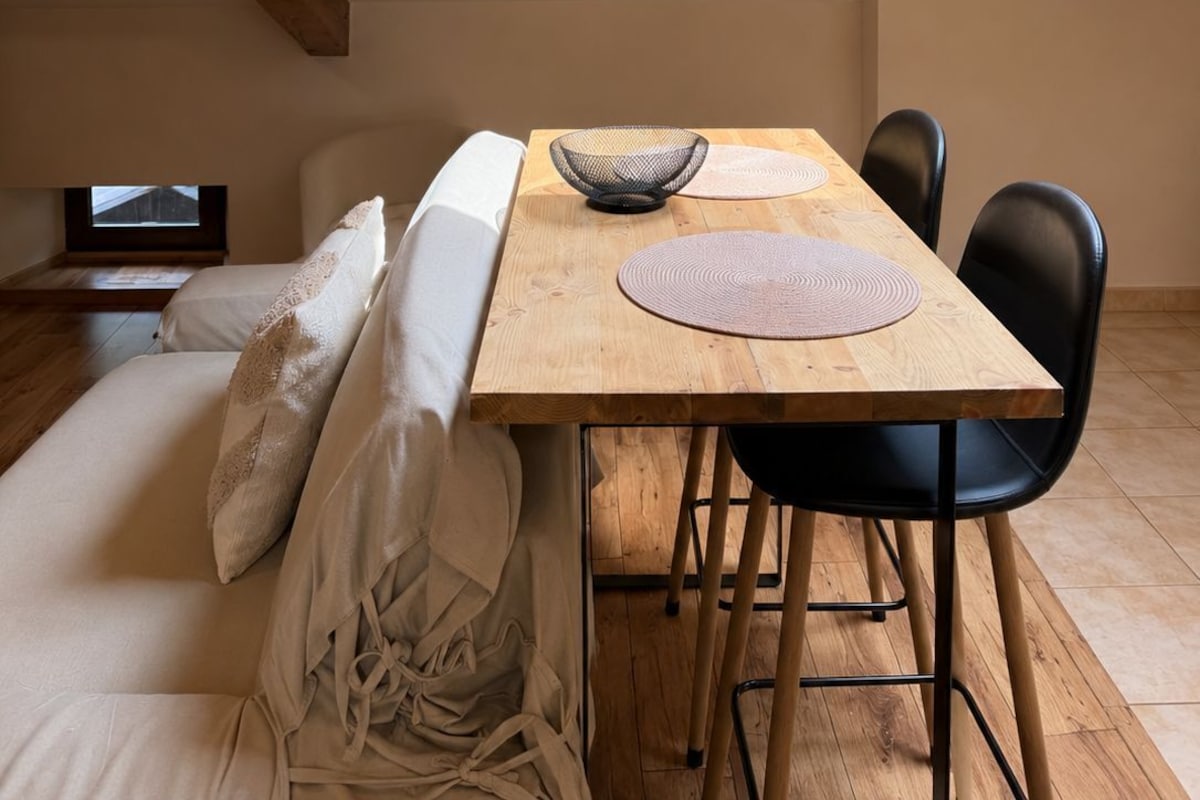 A cozy room with a wooden ceiling, a skylight, a wooden dining table with chairs, and a bed with pillows.