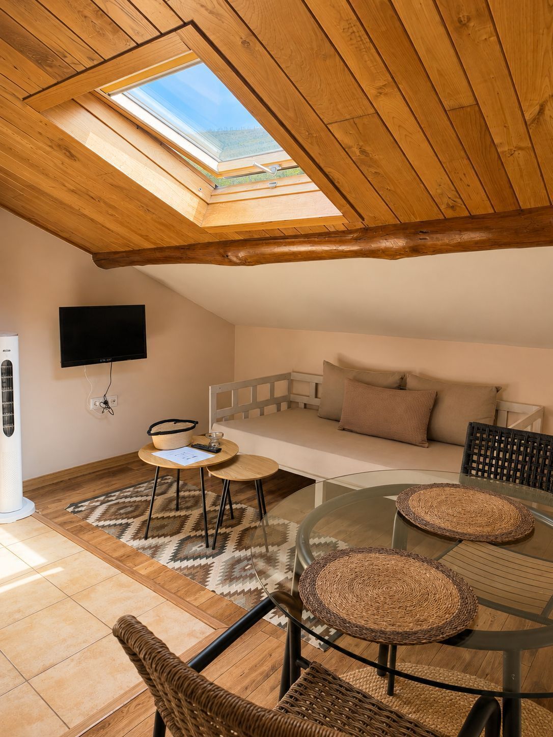Una habitación acogedora con techo inclinado, un sofá, una mesa de vidrio y un televisor montado en la pared.