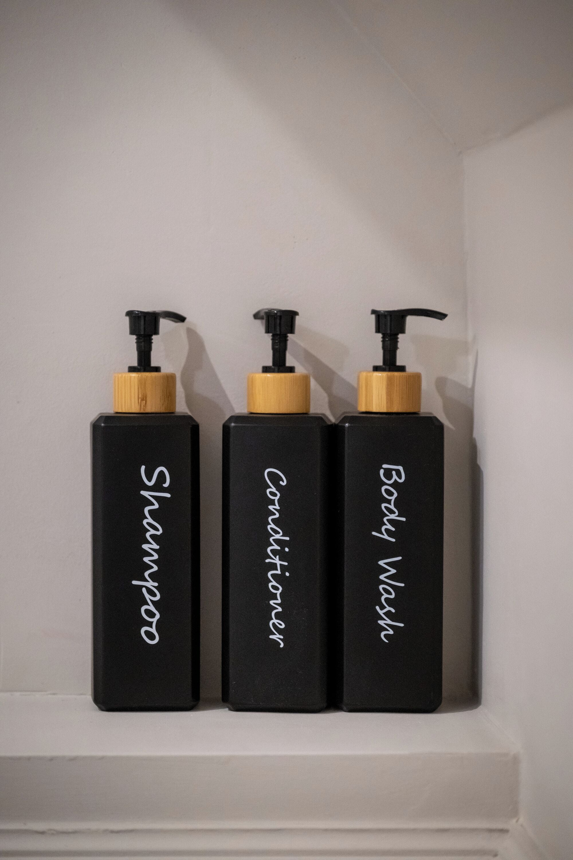 Trois bouteilles noires, munies de bouchons en bois, étiquetées « Shampoing », « Après-shampoing » et « Gel douche », sont posées sur une étagère blanche.