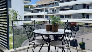 Ein Balkon mit rundem Tisch und Stühlen, Blumentöpfen und Blick auf ein mehrgeschossiges Gebäude.