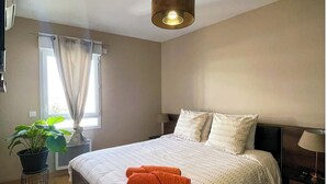 Ein ordentlich bezogenes Bett mit Kissen, einer Nachttischlampe und einer Hängeleuchte in einem Zimmer mit Fenster und einer Topfpflanze.