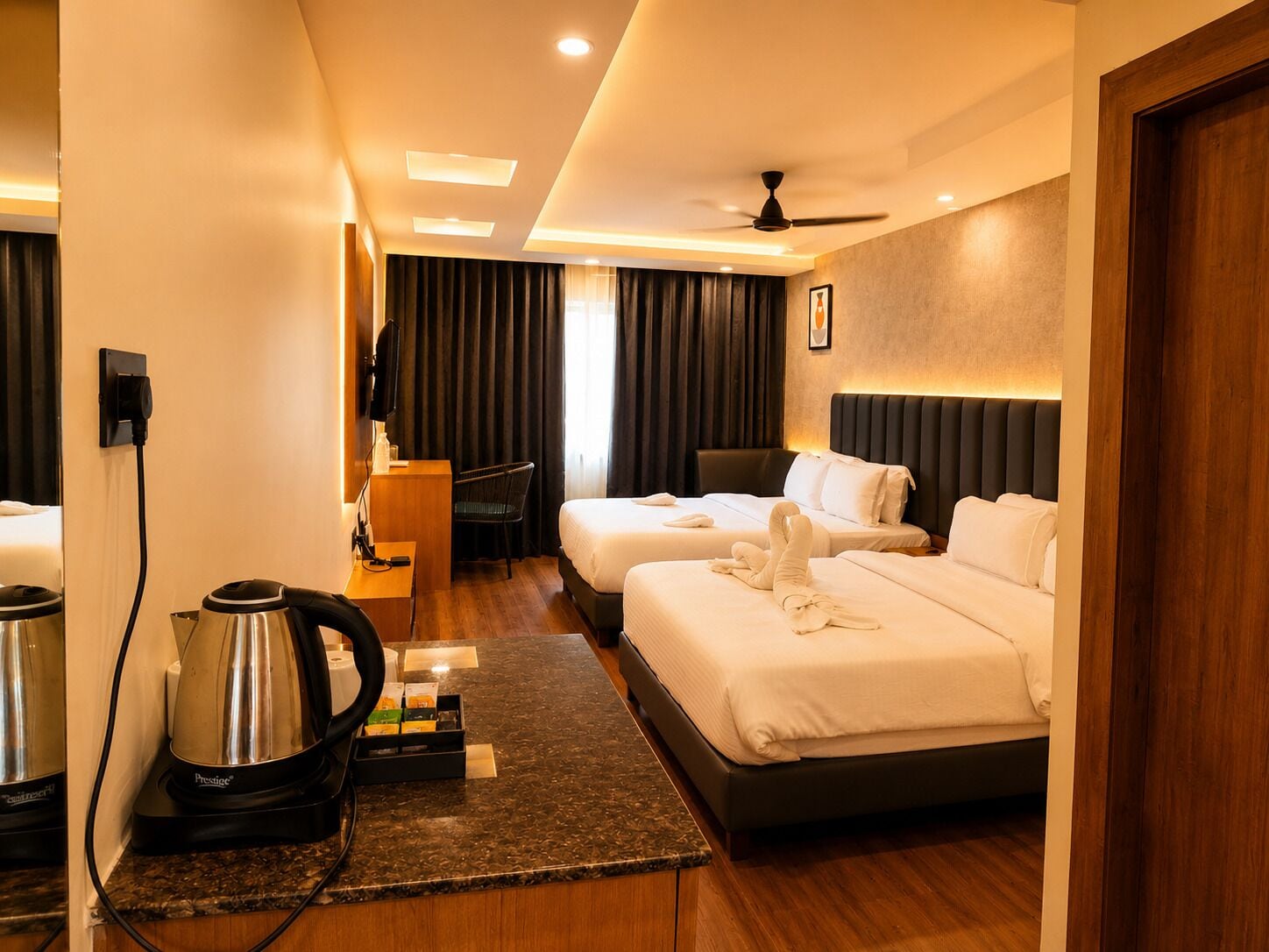 Une chambre d’hôtel avec deux lits, un bureau avec une bouilloire, une chaise, une télévision et un ventilateur de plafond.