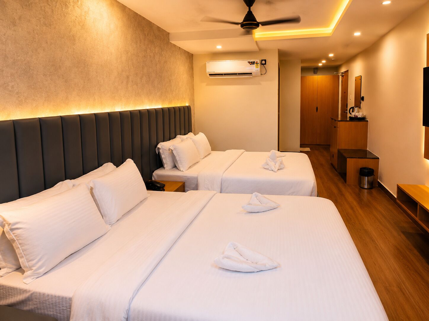 Habitación de hotel con dos camas, ventilador de techo, aire acondicionado, kitchenette pequeña y piso de madera.