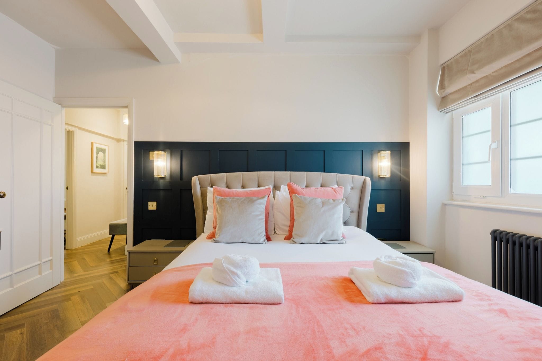 A hotel room with a large bed, two bedside tables, a nightstand, a window with curtains, and a wall-mounted light fixture.
