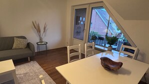 Un coin repas avec une table et des chaises blanches, une petite table d’appoint avec un vase, un canapé gris et une fenêtre donnant sur un bâtiment en briques.