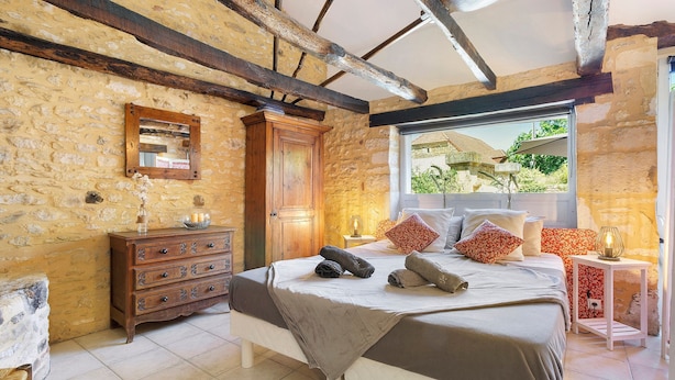 A bedroom with a stone wall, wooden beams, a bed with pillows, a dresser, and a window with a view. - Gîte "Le Four à Pain" – Charming Studio with Private Terrace in Périgord Noir (Paulin)
