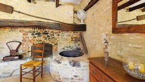 A rustic room with a stone wall, wooden beams, a stone stove, a wooden chair, a wooden dresser, and a mirror. - Gîte "Le Four à Pain" – Charming Studio with Private Terrace in Périgord Noir (Paulin)
