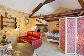 Una habitación rústica con vigas de madera a la vista, pared de piedra, sofá de cuero rojo, cama con cortinas florales y chimenea.