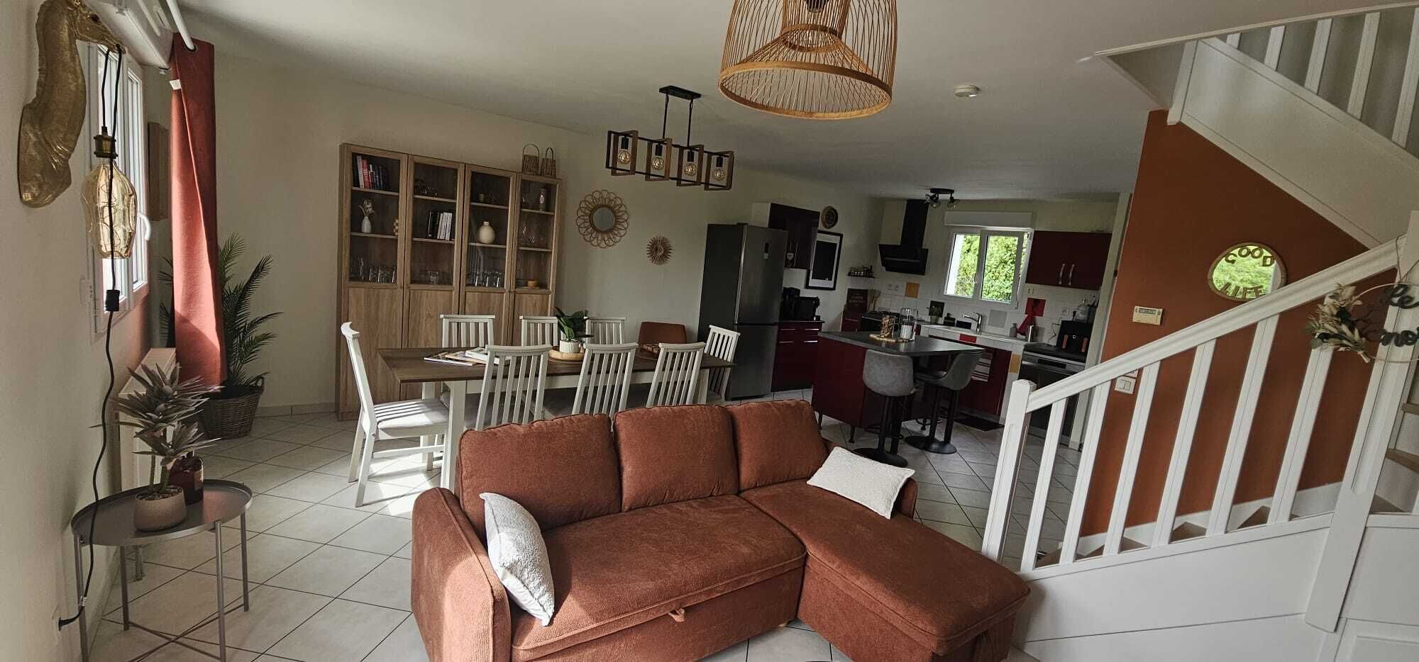 Een ruime woonkamer met een bank, eettafel en stoelen, een keuken op de achtergrond, en een trap naar een ander niveau.
