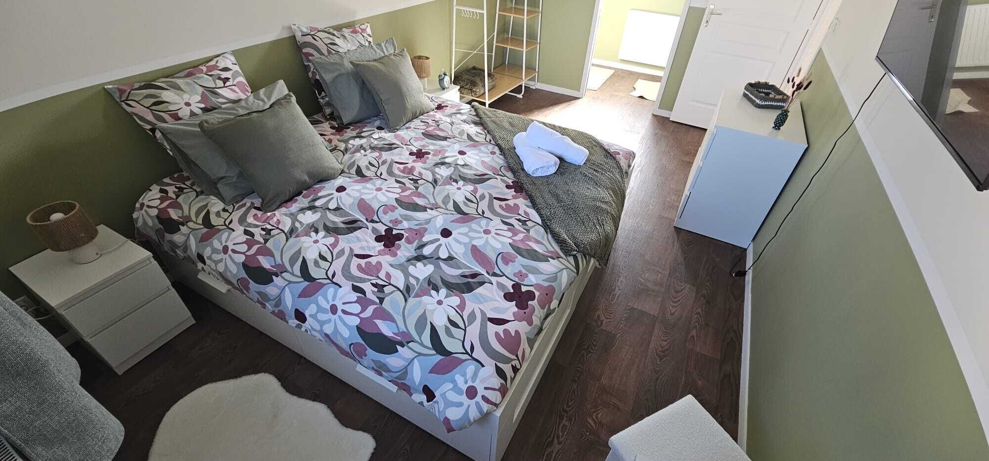 A bedroom with a bed, pillows, a nightstand, a wardrobe, and a small table with a lamp.