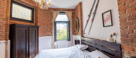A bedroom with a brick wall, a bed, a chandelier, a window, and skis on the wall.