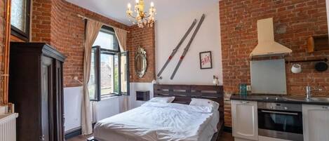 Un dormitorio con cama, candelabro, chimenea, zona de cocina y pared de ladrillo.