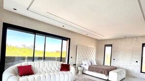 Een moderne woonkamer met een hoekbank, een bed en een groot raam met uitzicht op groen.