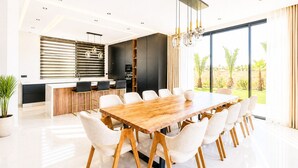 Een moderne keuken met een centraal eiland, een eetgedeelte met een grote houten tafel en witte stoelen, en een groot raam met uitzicht op groen.