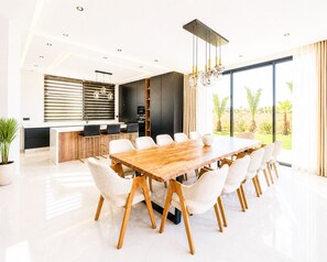 Eine moderne Küche mit einer zentralen Kochinsel, einem Essbereich mit einem großen Holztisch und weißen Stühlen sowie einem großen Fenster mit Blick auf Grünflächen.