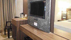 Une chambre d’hôtel avec un téléviseur fixé au mur en marbre, un bureau en bois avec une chaise, un lit avec une tête de lit et une table de chevet avec une lampe.