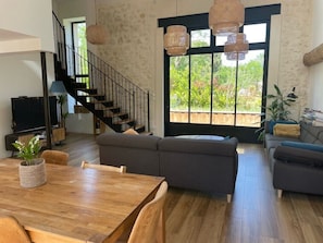 A spacious living area with a wooden dining table, sofas, a staircase, and large windows with a view of greenery. - Renovated 17th-century wine storehouse, 4 km from Bordeaux-Lacanau cycle path. (Le Taillan-Médoc)