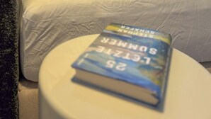 Una cama con ropa de cama blanca y una almohada amarilla, un libro sobre una mesita de noche y vistas de vegetación a través de una ventana.